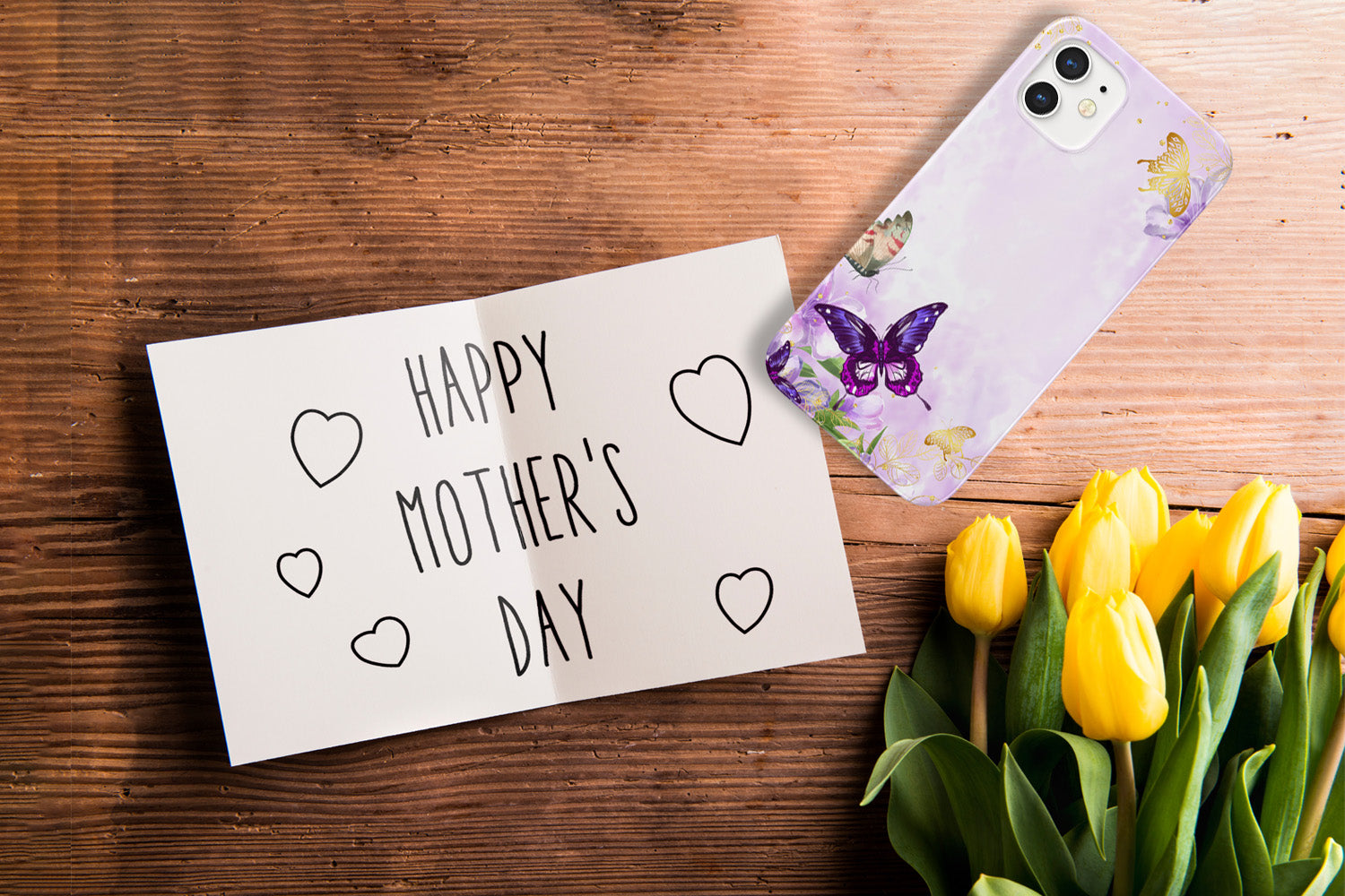 Table with a bouquet of yellow flowers. A note that reads Happy Mother's Day with hand drawn hearts. An iPhone with a Gilded Flutters Butterfly phone case.