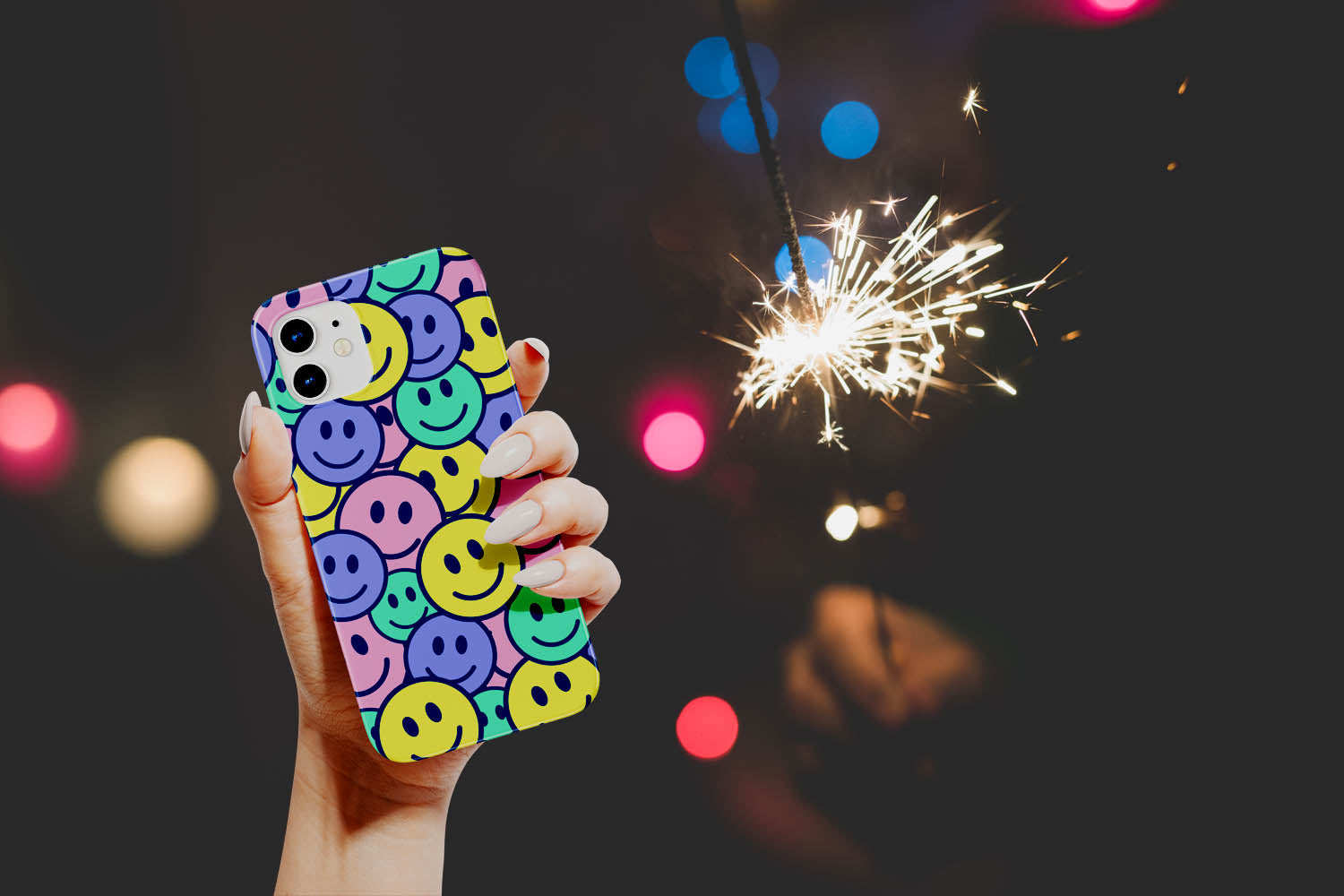 Nighttime photo with fireworks in the background and a close up of a hand holding a sparkler and another hand holding a Groovy Smiles Smiley Face Phone Case